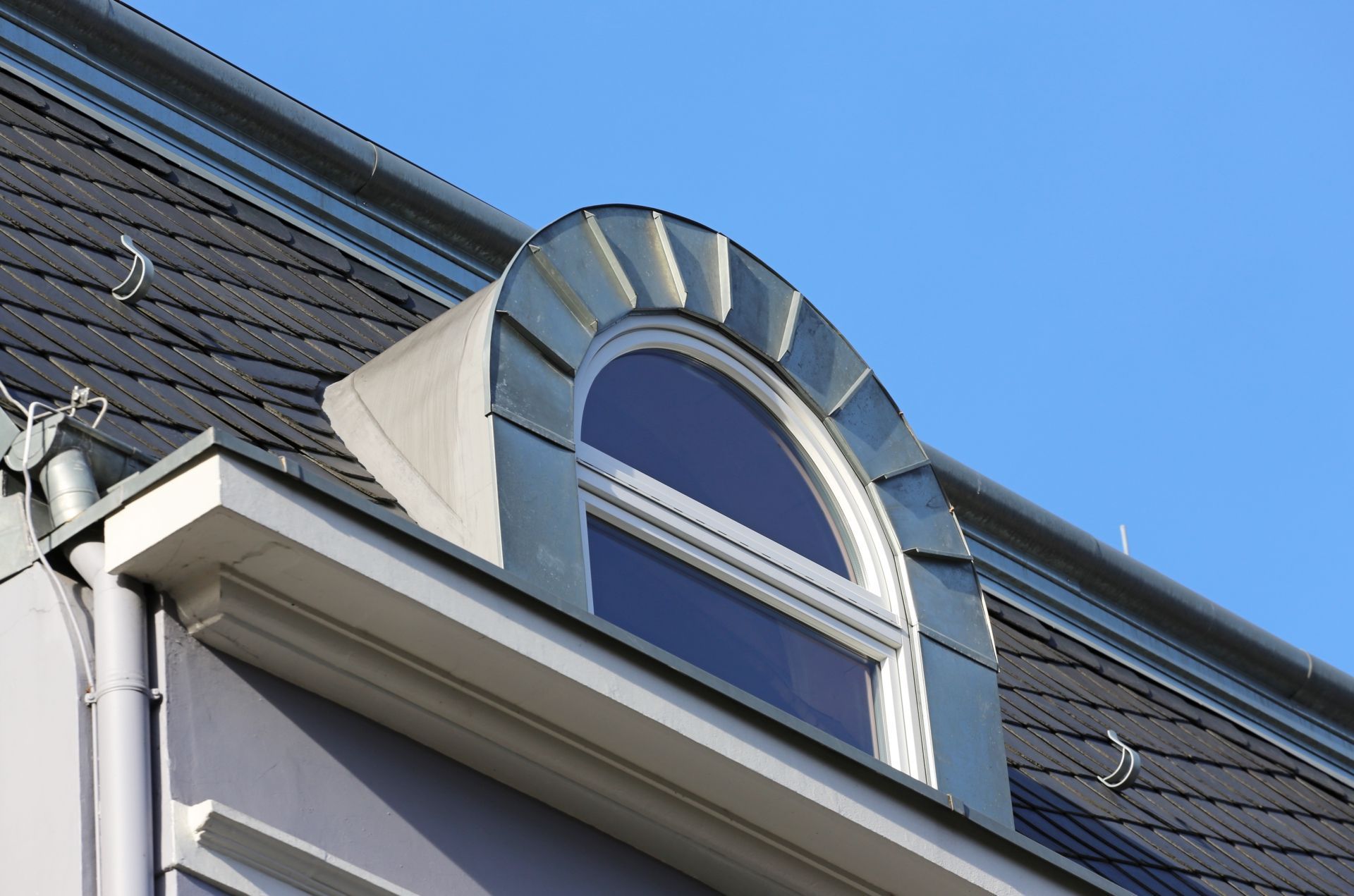 Dachgaubenfenster mit bogenförmigem, blauem Zierrahmen und weißer Verkleidung an der Dachlinie eines Gebäudes