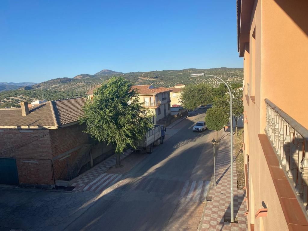 Vista de la calle en una ciudad soleada con edificios, árboles, un vehículo y colinas distantes bajo un cielo azul.