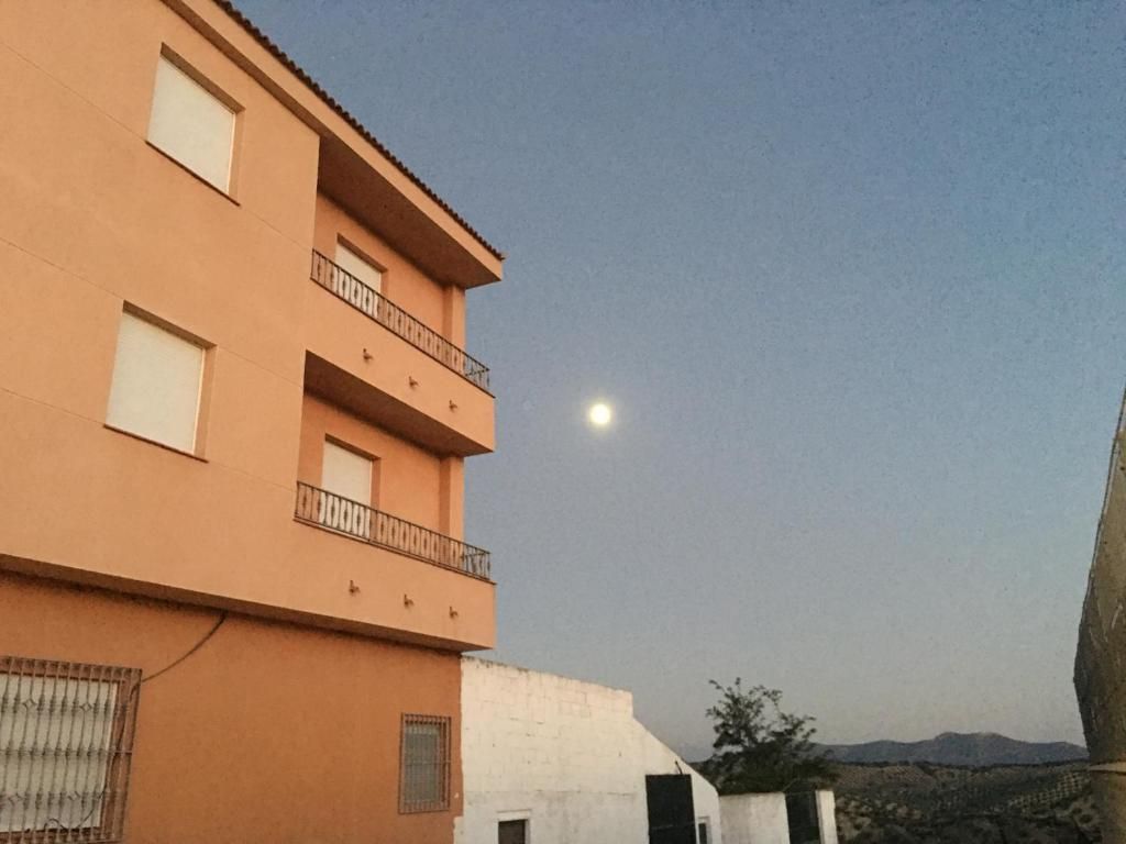 Edificio naranja con balcones, luna llena en un cielo azul pálido.