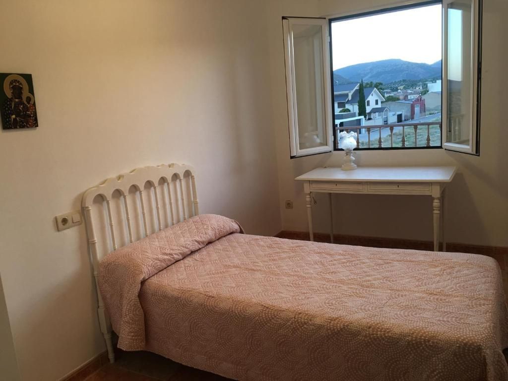 Dormitorio con cama blanca, pequeño escritorio y ventana abierta con vistas a un pueblo.