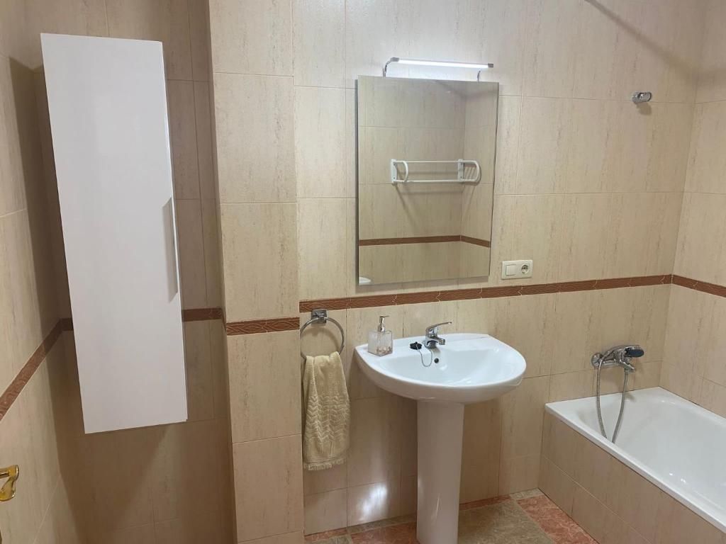Baño con lavabo de pedestal, espejo, mueble y bañera. Paredes de azulejos beige con detalles marrones.