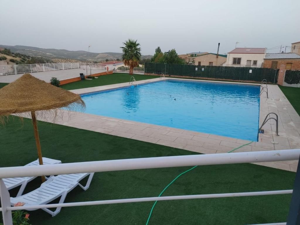 Piscina con césped, tumbonas y sombrilla. Al fondo se ven edificios y una ladera.