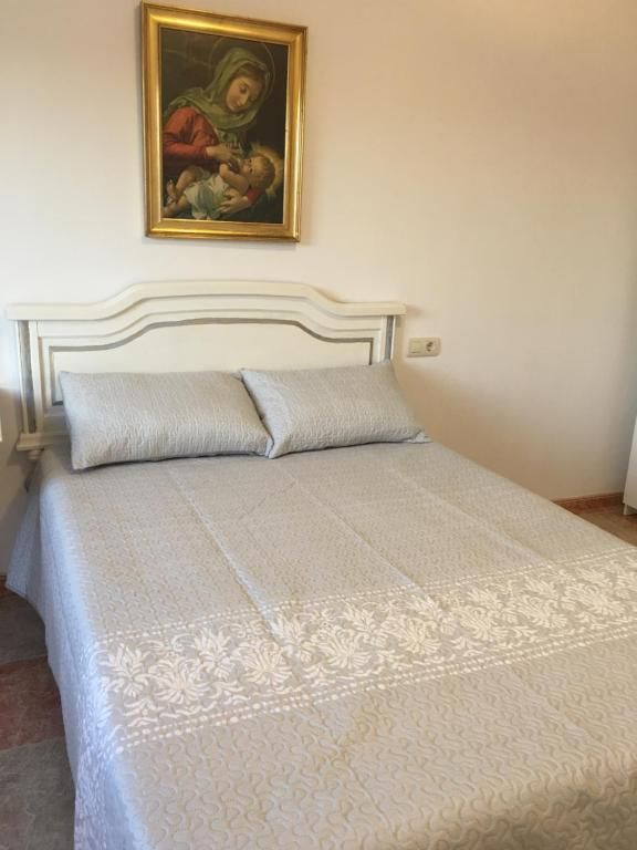 Dormitorio con una cama, dos almohadas, colcha de color claro y obra de arte religioso enmarcada sobre la cabecera.