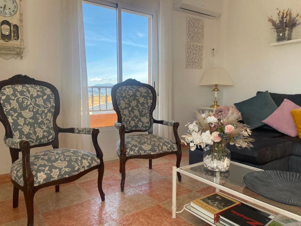 Dos sillas ornamentadas frente a una ventana, sala de estar con sofá, mesa de café y ramo de flores.