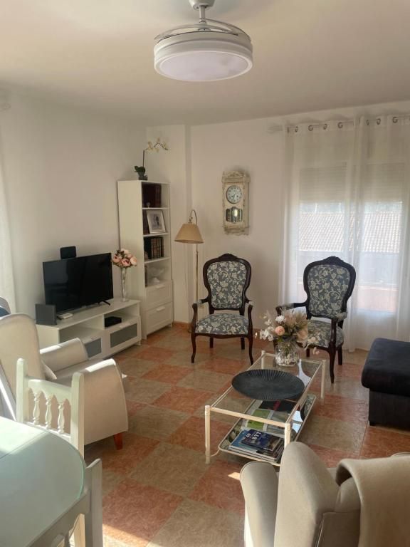 Sala de estar con muebles blancos, sillas con motivos florales, televisor y suelo de baldosas estampadas. La luz del sol entra a raudales por una ventana.