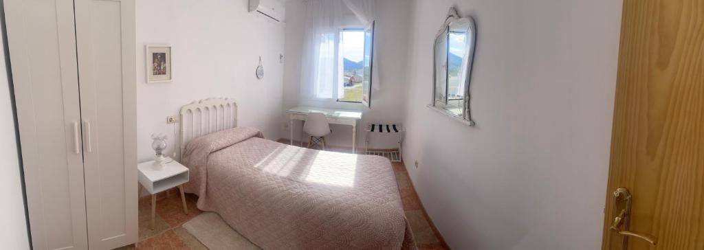 Dormitorio con paredes blancas, una cama, un pequeño escritorio y un espejo decorativo.