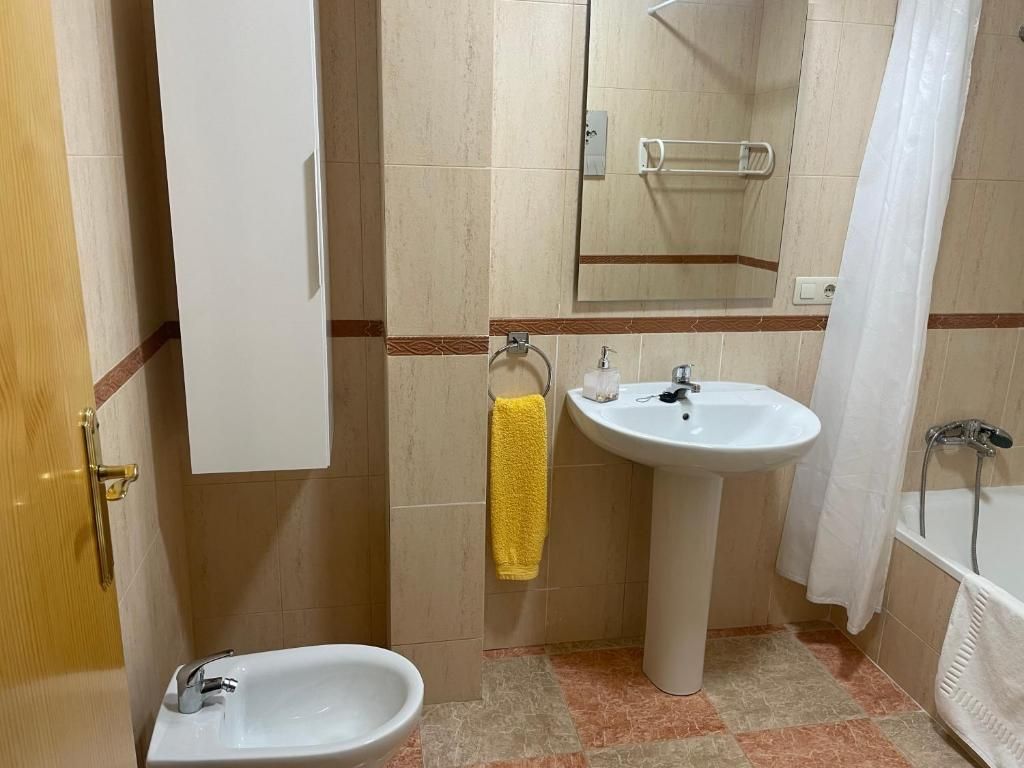 Baño con lavabo, bidé, mueble y bañera blancos. Se ven azulejos beige y una toalla amarilla.