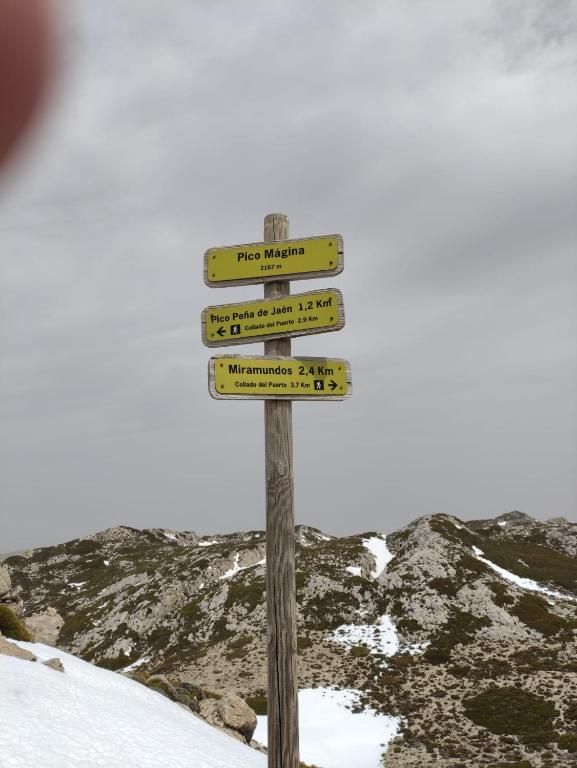 Señal de sendero de madera con marcadores amarillos contra un fondo de montañas nevadas, que apunta a Pico Magnu y otros lugares.