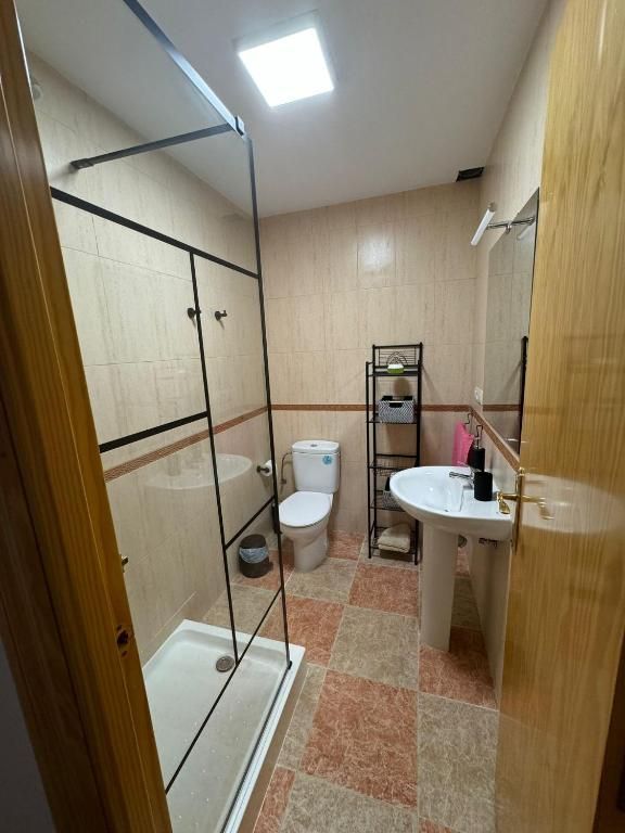 Baño con ducha, inodoro, lavabo y estantes; paredes de color beige claro, suelo de baldosas de color marrón rojizo y tragaluz.