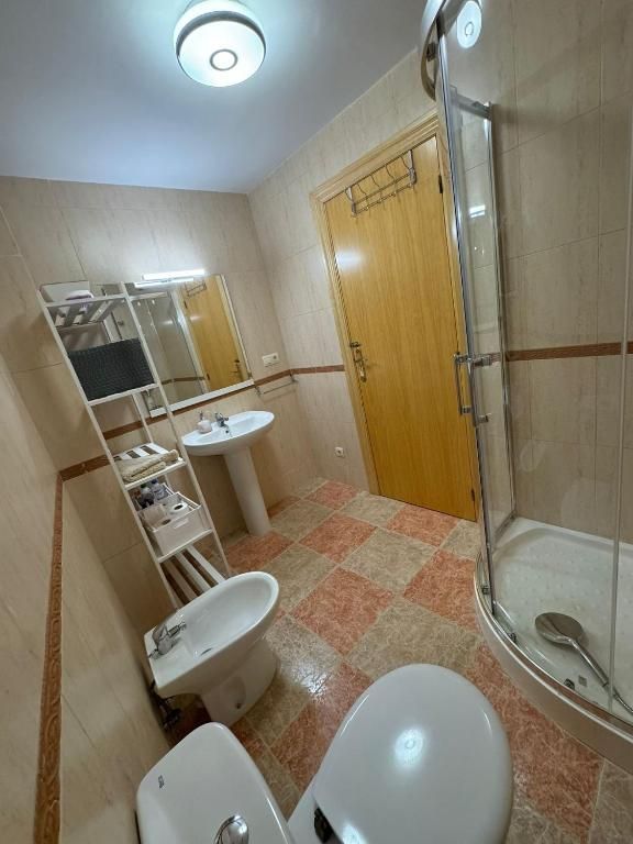 Baño con inodoro, bidé, lavabo, ducha y estantería. Paredes beige, suelo de baldosas, puerta de madera y luz cenital.