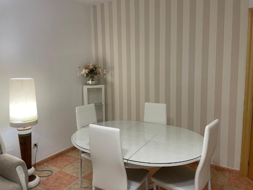 Comedor con mesa redonda blanca, sillas blancas, pared rayada y lámpara de pie.