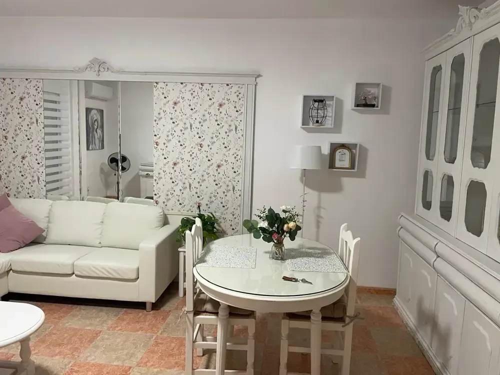 Sala de estar blanca con sofá, mesa y armario. Se ven un espejo y persianas estampadas.