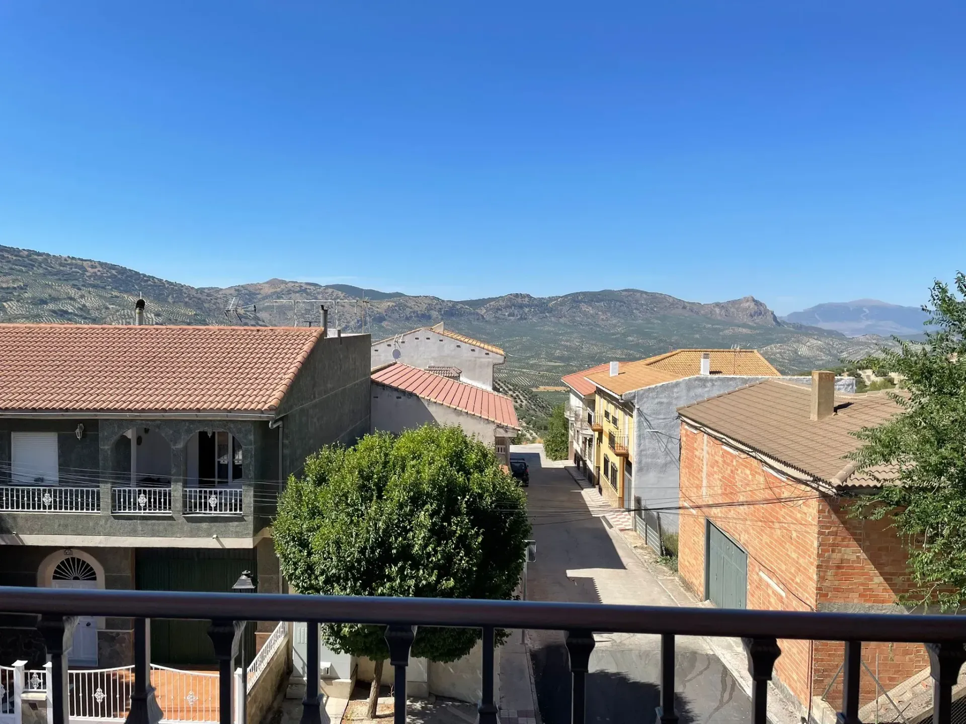 Vista de una calle bordeada de edificios y techos de tejas rojas, montañas al fondo, bajo un cielo azul.