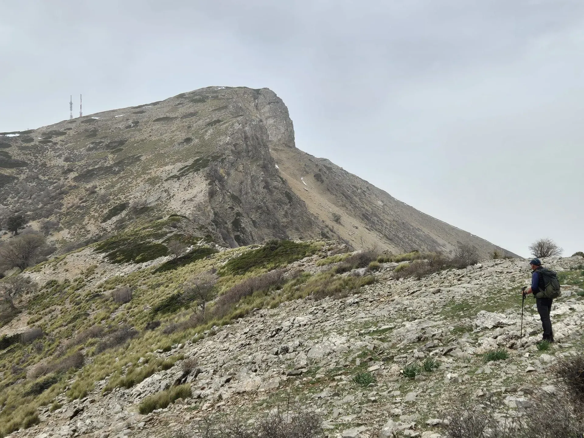 Una persona camina por un sendero rocoso en la montaña hacia un pico con antenas, bajo un cielo nublado.