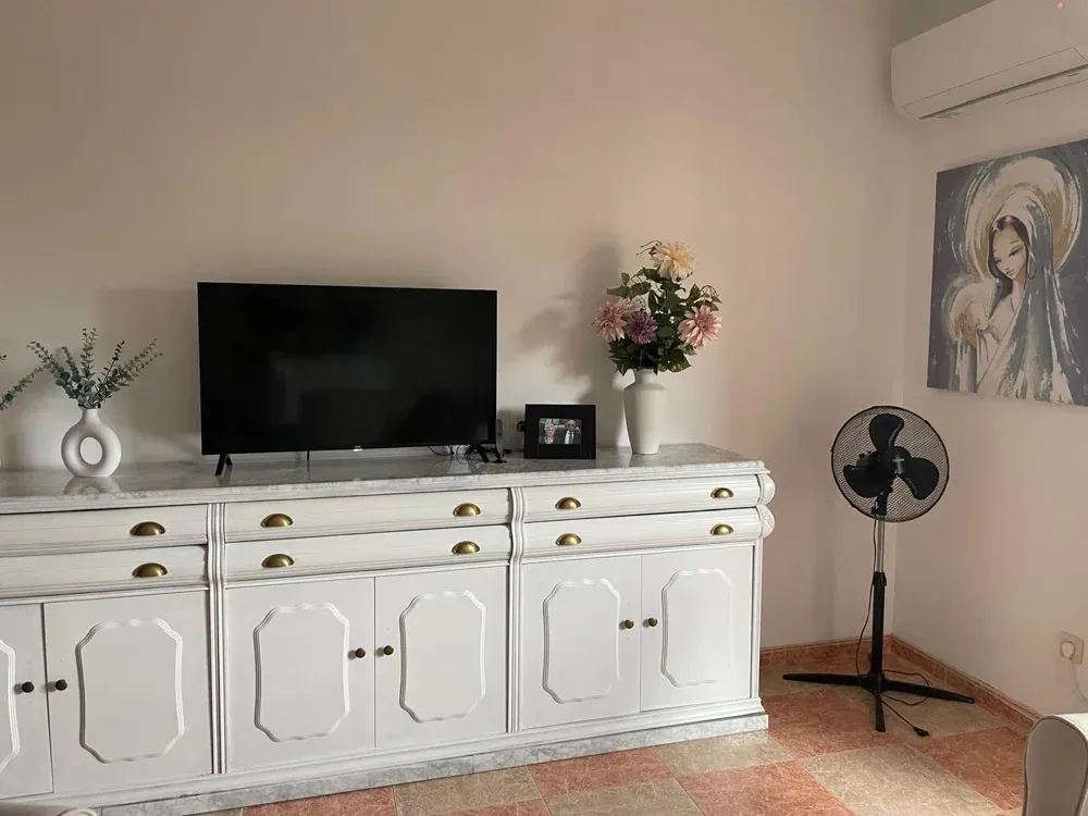 Mueble blanco con TV, flores y un ventilador de pie en una habitación con un cuadro.