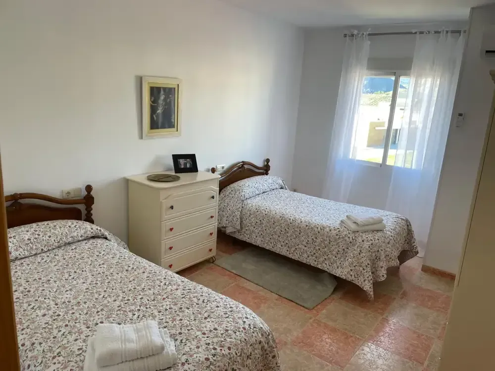 Dos camas individuales en una habitación luminosa con cómoda blanca, ventana y ropa de cama con motivos florales.