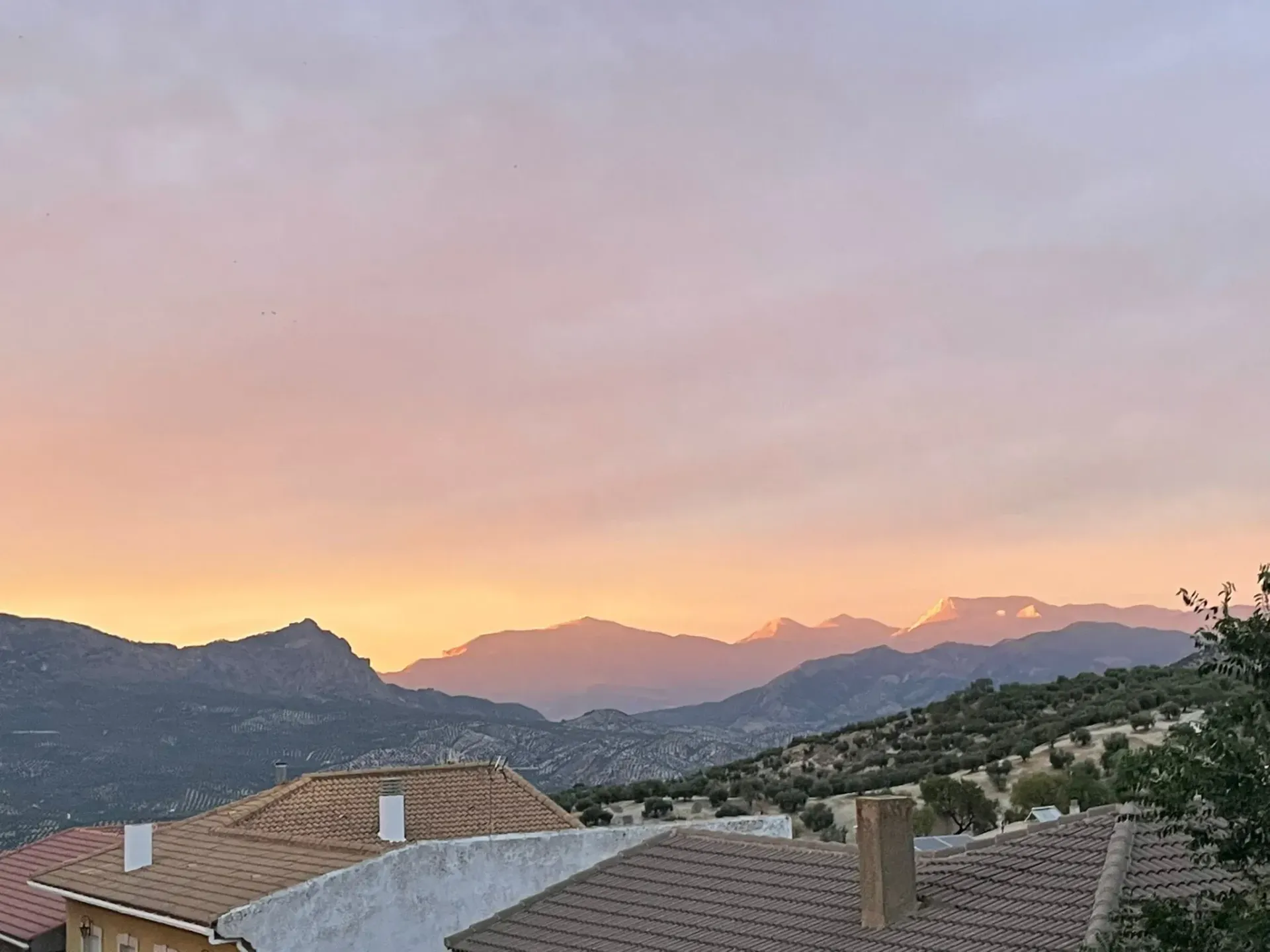 Los tejados se asoman a una cordillera al atardecer. El cielo presenta un degradado naranja y rosa.