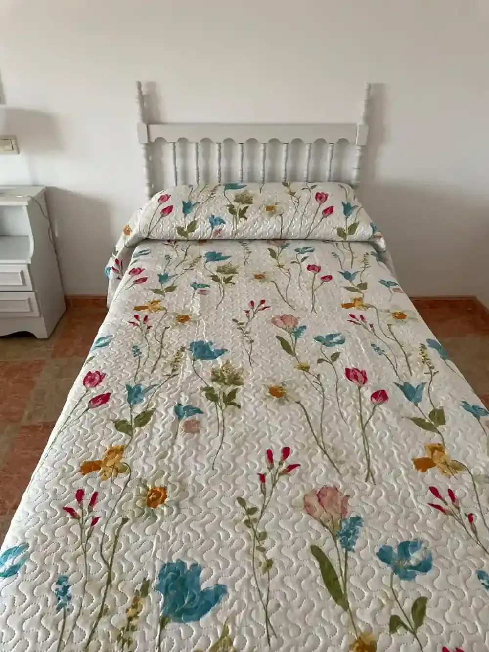 Una cama individual con edredón con estampado floral y cabecero blanco en una habitación llena de luz.