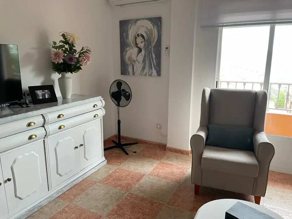 Sala: mueble blanco, arreglo floral, ventilador, sillón, cuadro, piso de baldosas, ventana.