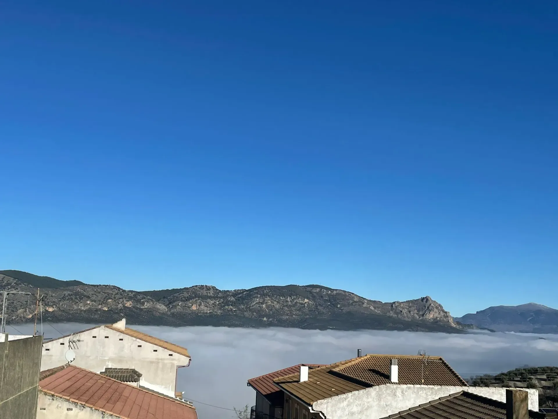 Casas con techos de tejas bajo un mar de nubes, montañas al fondo y un cielo azul claro arriba.