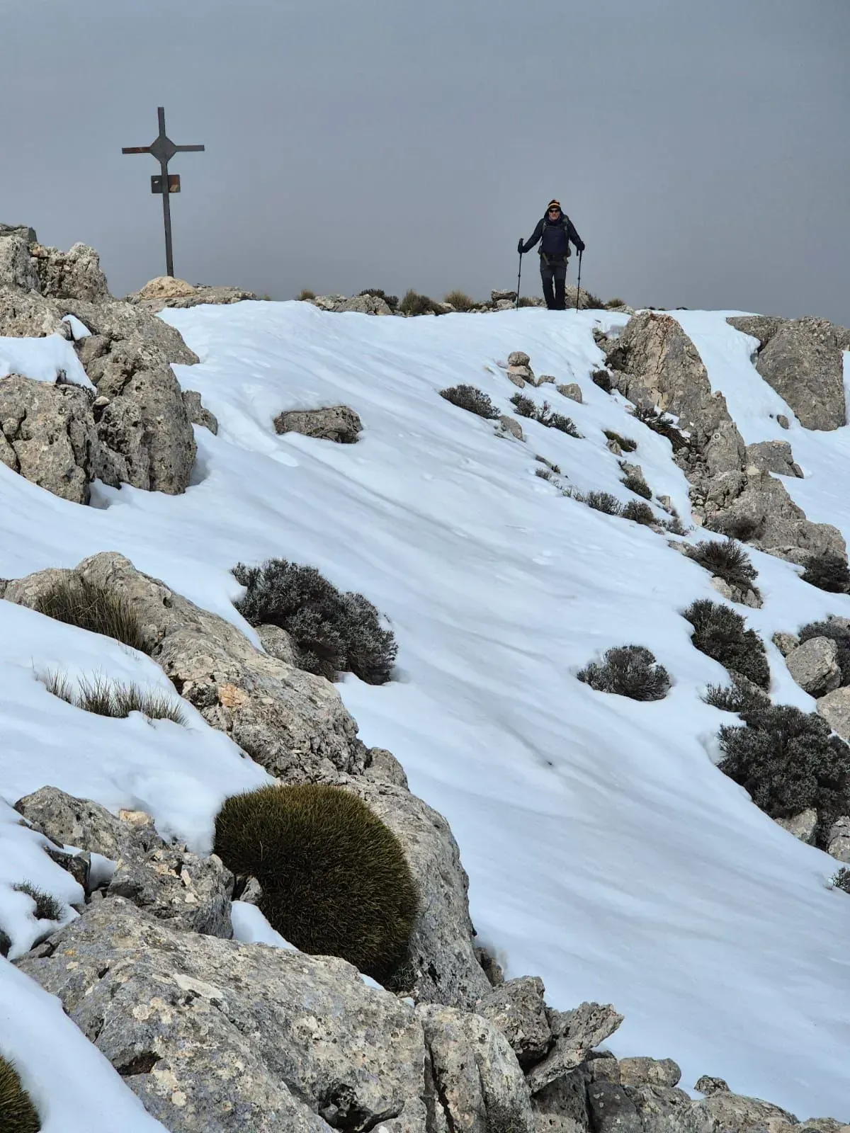 Una persona con bastones de trekking se encuentra cerca de una cruz en la cima de una montaña nevada.