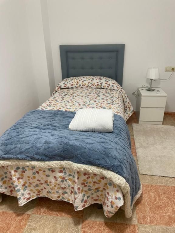 Cama individual con edredón y cabecero de color azul, mesita de noche y lámpara pequeña.