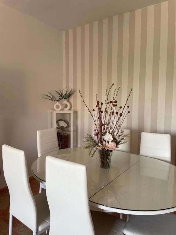 Comedor con sillas blancas alrededor de una mesa con tapa de cristal, centrada por un arreglo floral, sobre un papel tapiz a rayas.
