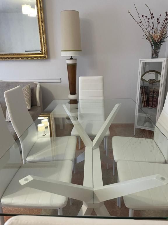 Mesa de comedor de vidrio con sillas blancas, una lámpara y una vitrina contra una pared neutra.