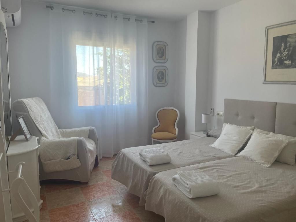 Dormitorio con dos camas individuales, una silla y una ventana con cortinas transparentes.