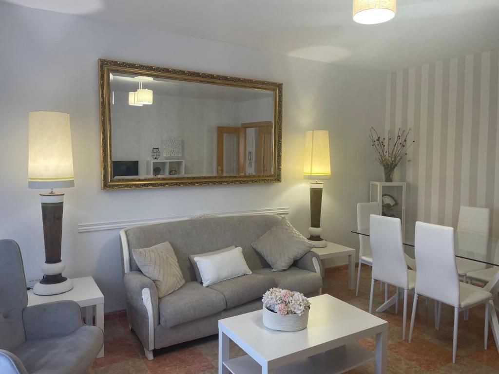 Sala de estar con sofá, espejo, lámparas, mesa, mesa de comedor y sillas. Tonos beige y blanco.