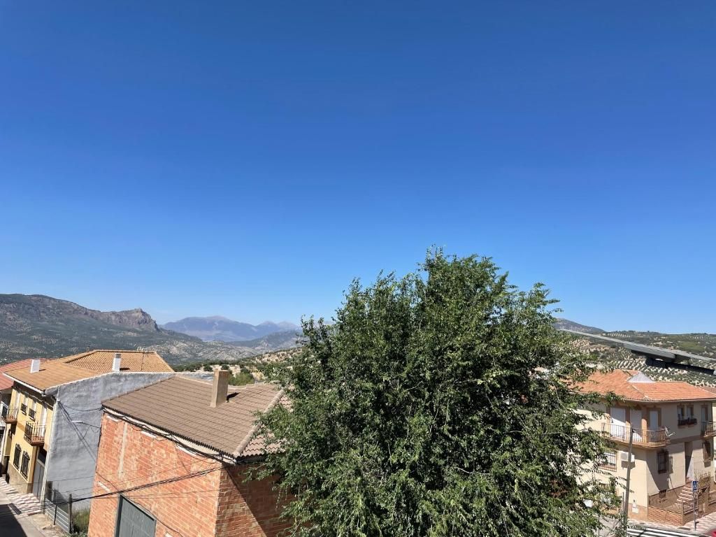 Paisaje urbano bajo un cielo azul brillante, con edificios, un árbol y montañas en la distancia.