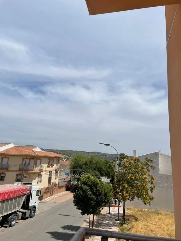 Escena callejera con edificios, árboles, un camión y un cielo nublado visto desde un balcón.