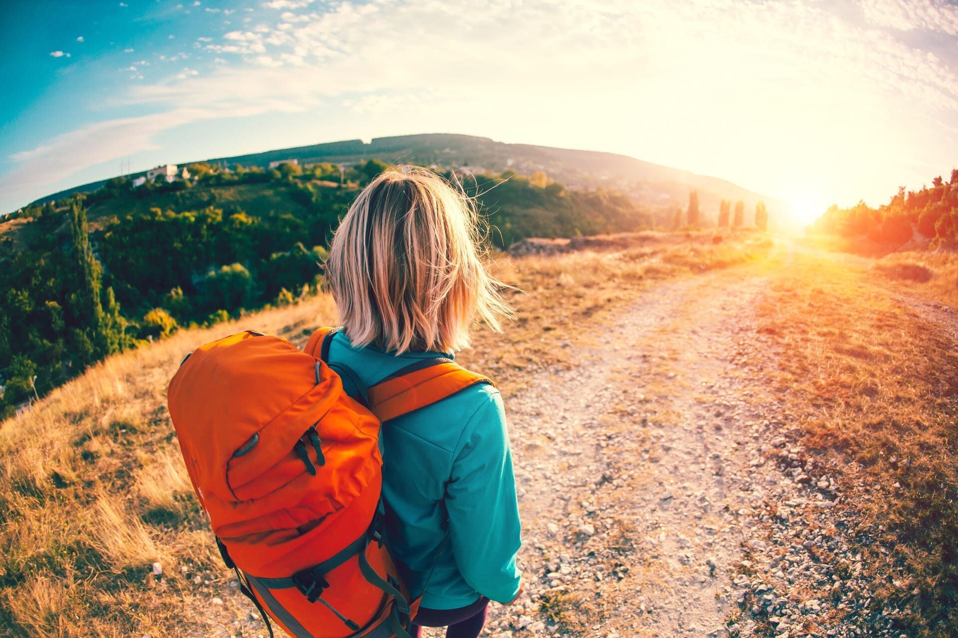Mujer con mochila naranja caminando por un sendero soleado hacia el amanecer.