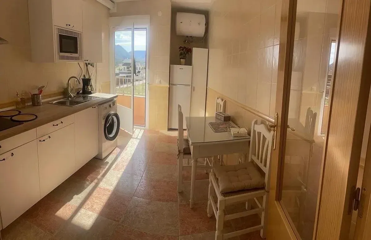 Cocina con gabinetes blancos, electrodomésticos y una pequeña mesa de comedor. La luz del sol entra a raudales por la ventana.