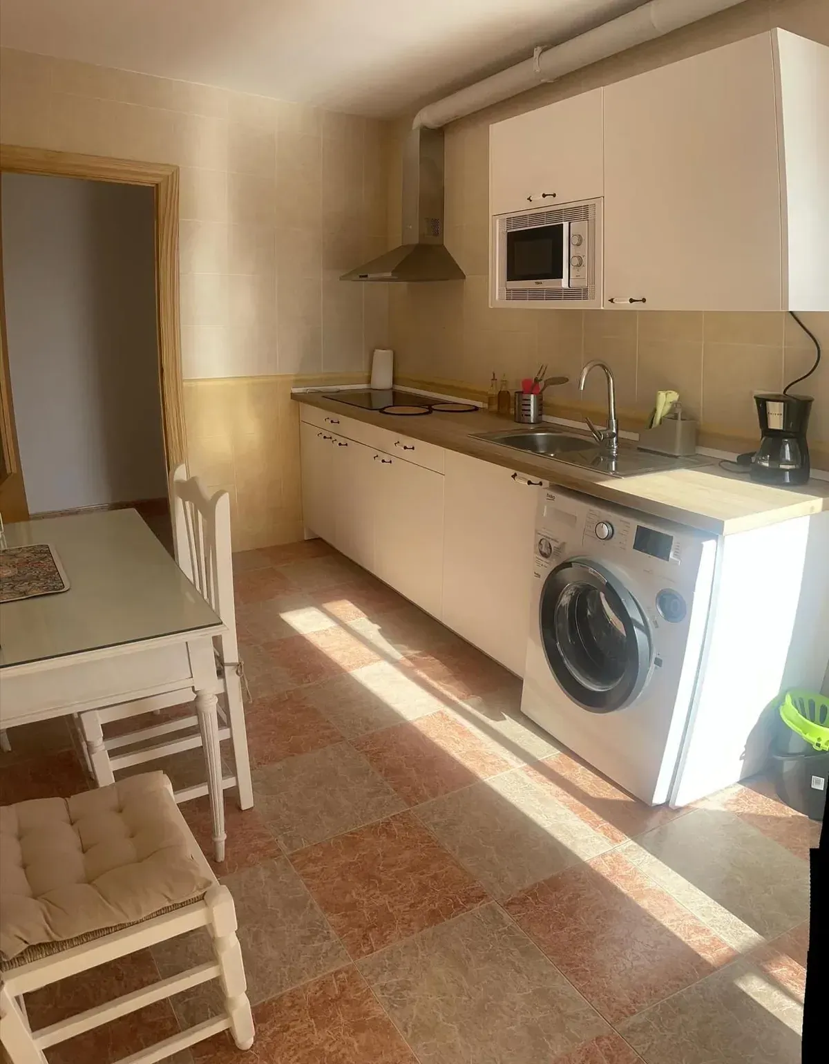 Cocina con muebles blancos, electrodomésticos y mesa con sillas. Suelo de baldosas color marrón anaranjado.