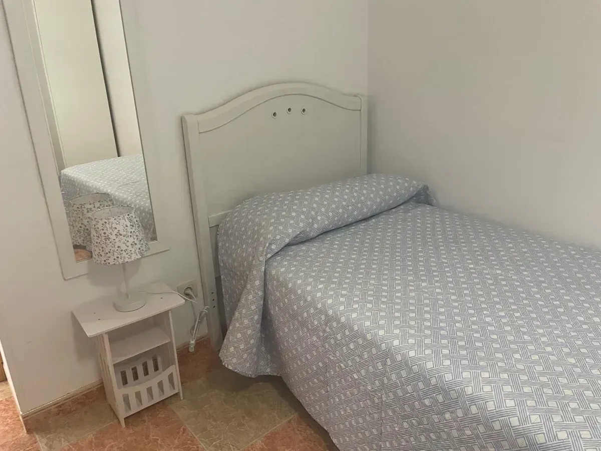 Un dormitorio pequeño con una cama individual cubierta con una colcha estampada, una mesita de noche con una lámpara y un espejo.