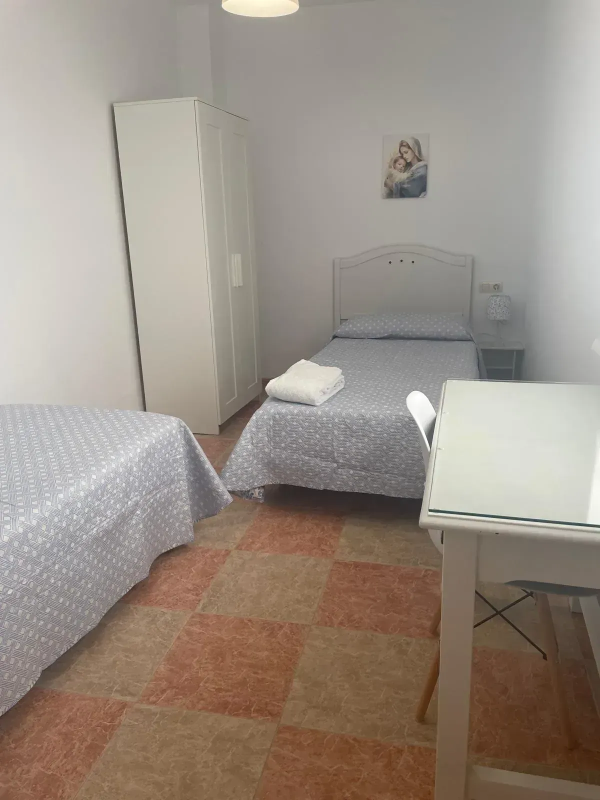 Dos camas individuales en una habitación con un armario blanco y un pequeño escritorio; ropa de cama estampada, suelo de baldosas.