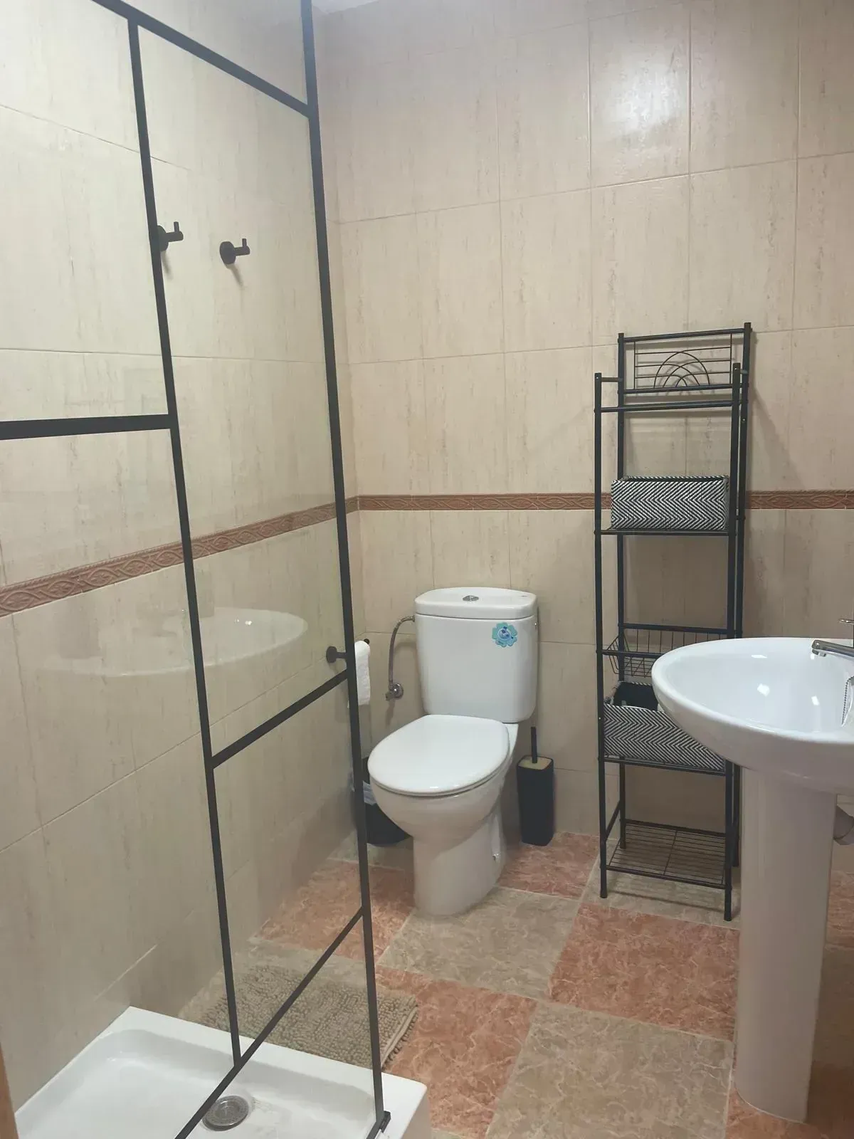 Baño con inodoro, lavabo, ducha y estantes; suelo y paredes de baldosas de color marrón y tostado.
