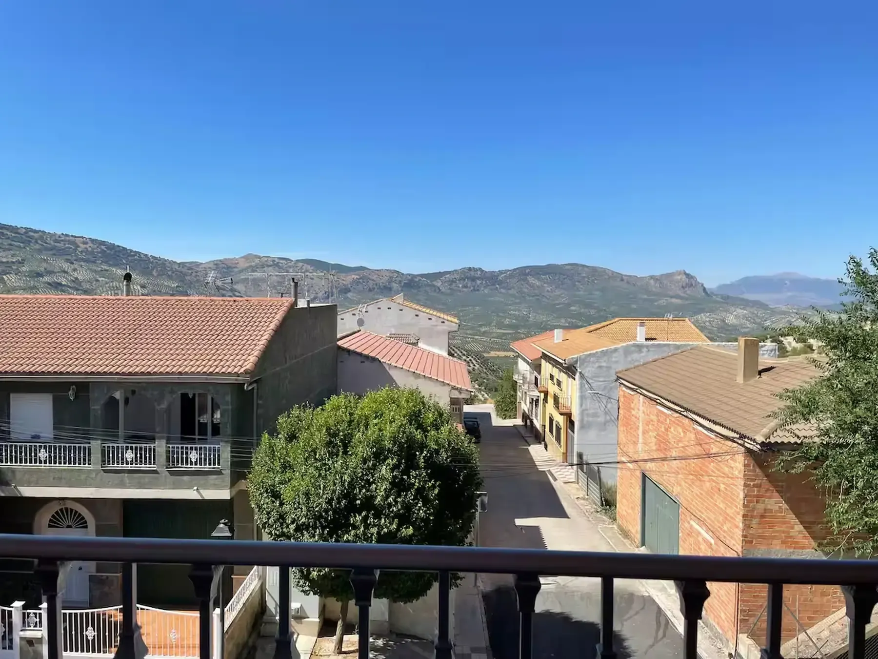Vista de una calle de un pueblo, con casas y montañas al fondo bajo un cielo azul.