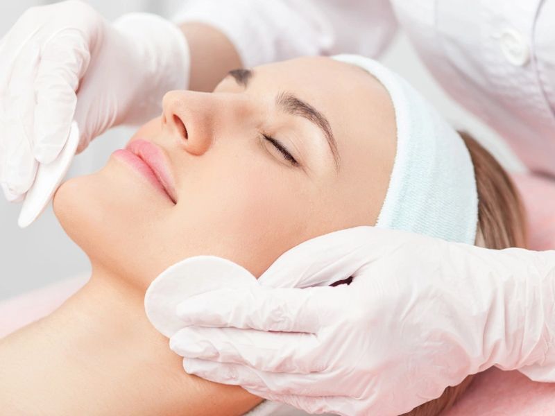 Manos enguantadas aplicando un tratamiento facial a una clienta relajada con una diadema blanca.