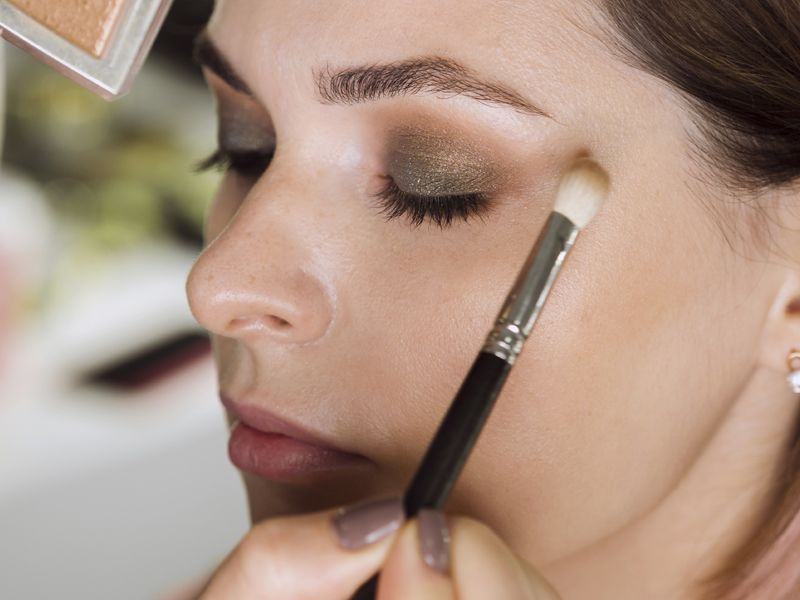 Primer plano de una mujer aplicándose maquillaje de ojos con una brocha, con los ojos cerrados, en un entorno con iluminación tenue.