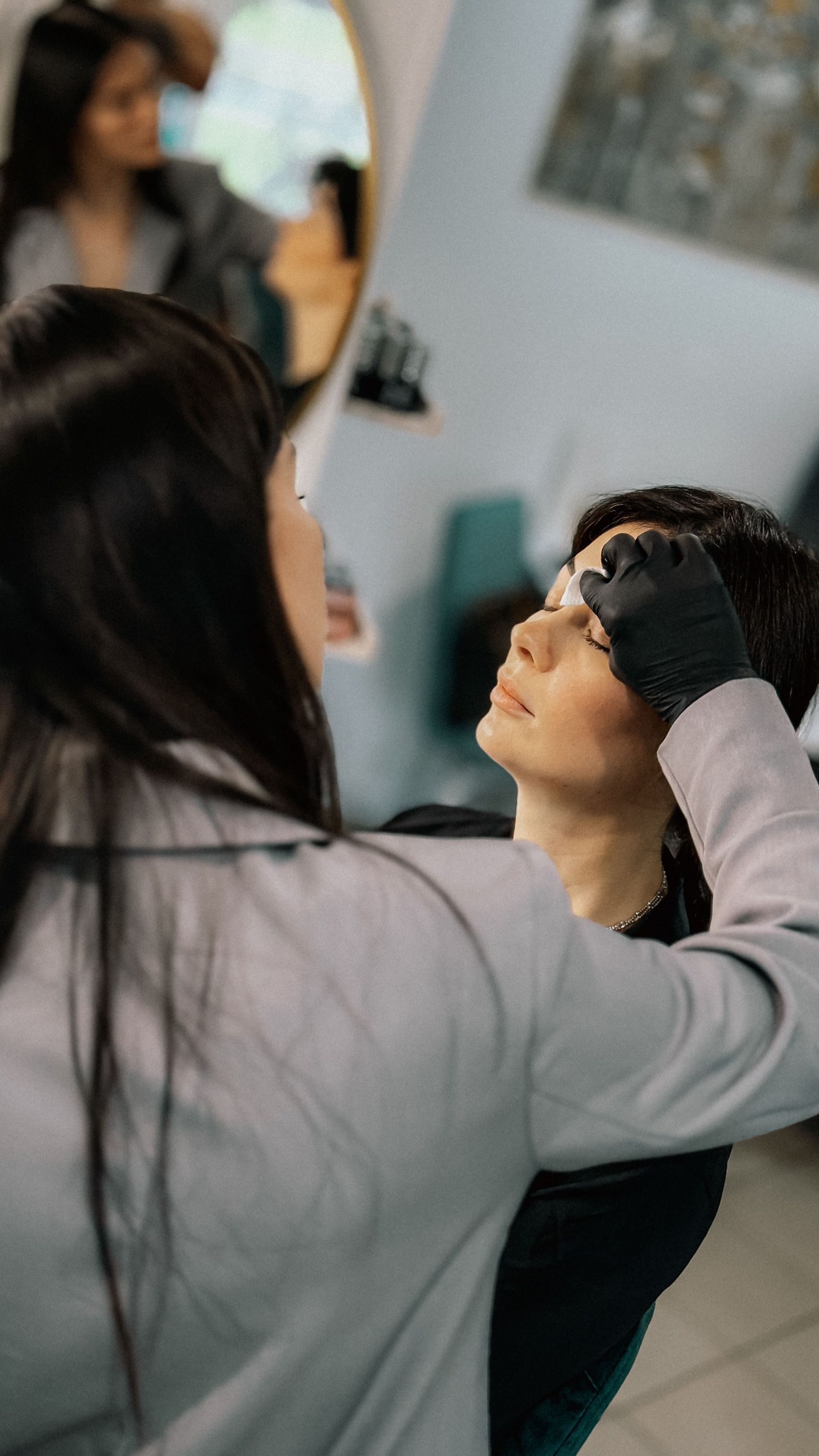 Una persona con guantes negros le arregla el rostro a una persona sentada durante un tratamiento de maquillaje o belleza.