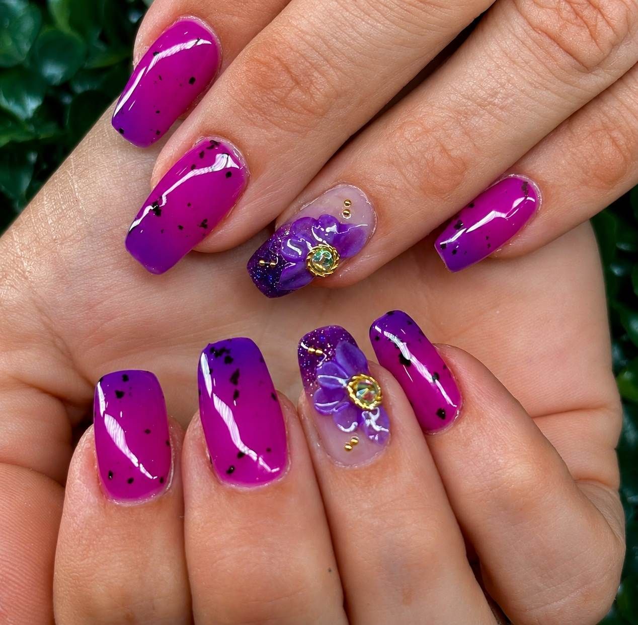 Manicura con purpurina morada y diseño floral en uñas largas y brillantes.