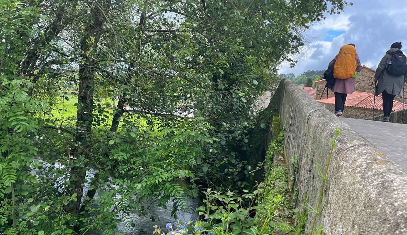 Dos personas con mochilas caminan por un puente sobre un río.