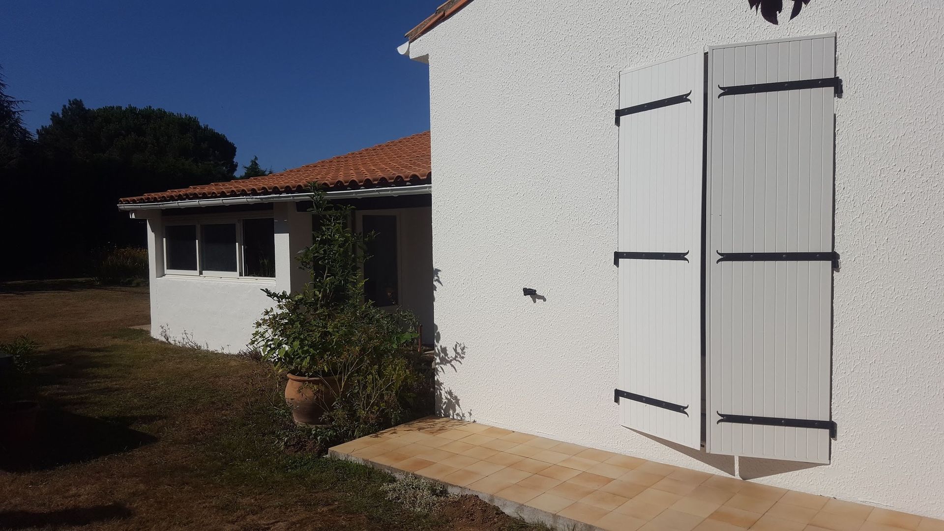 Maison blanche avec volets, toit de tuiles rouges et allée marron par une journée ensoleillée.