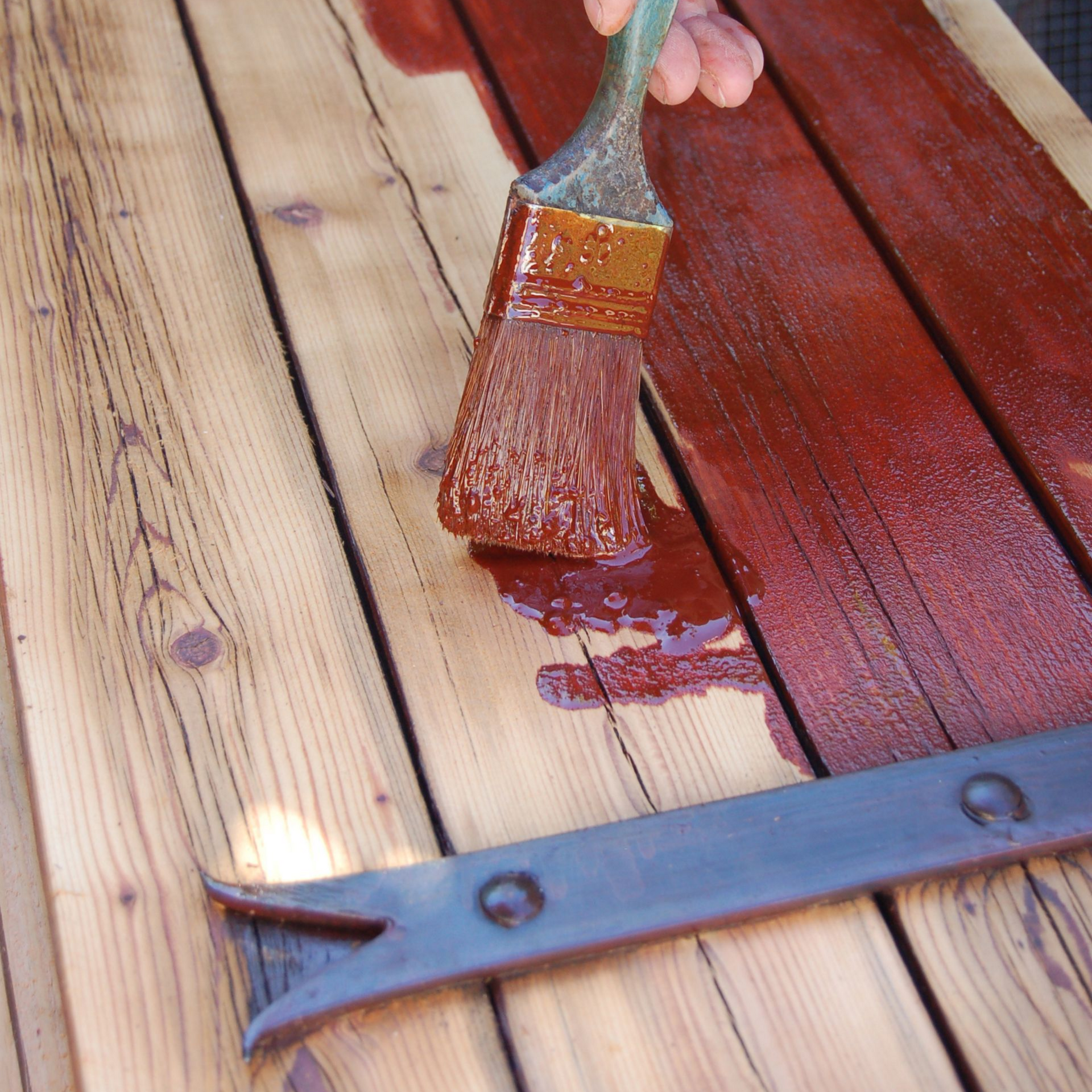 Une personne utilise un pinceau pour appliquer une teinture rouge sur des planches, avec une charnière métallique visible.
