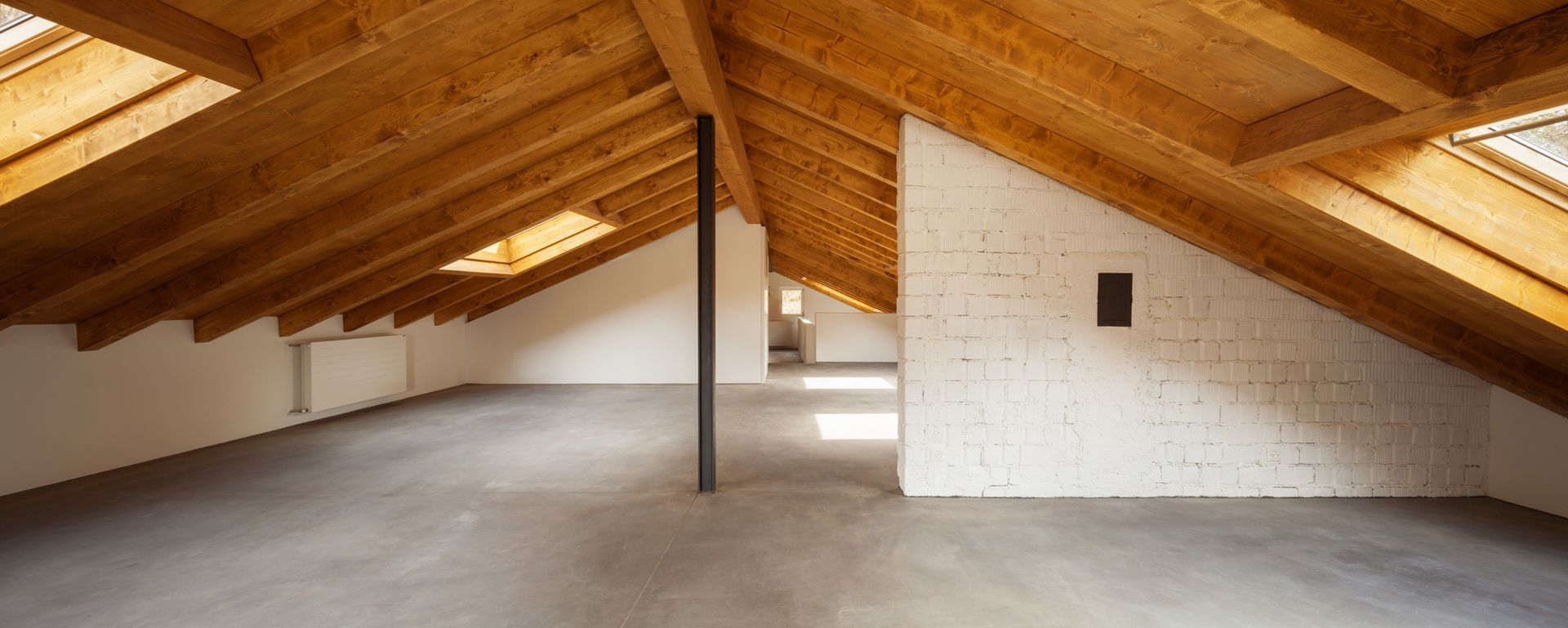 Un grenier vide avec des poutres en bois et un sol en béton ; la lumière naturelle coule des puits de lumière.