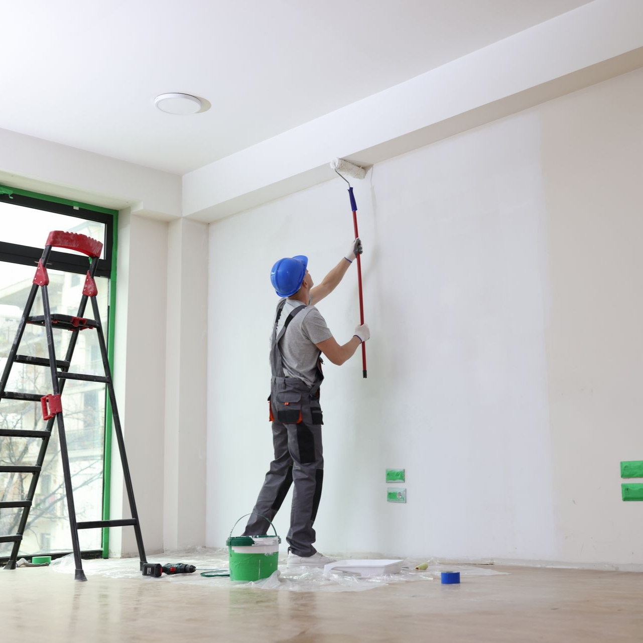 Un peintre portant un casque bleu et une salopette peint un mur blanc avec un rouleau, debout sur le sol.