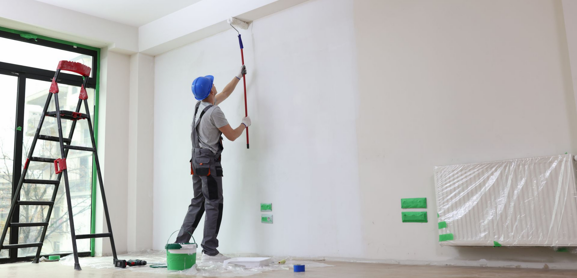 Un peintre en salopette et casque bleu peint un mur en blanc avec un rouleau.