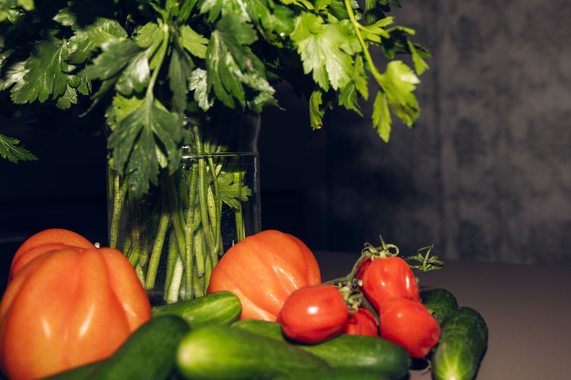 Des tomates, des concombres et du persil, disposés dans un vase en verre transparent, sont posés sur un comptoir.